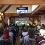 A large group of people listens to a presentation on turtle rehabilitation with photos of turtles on a large screen.