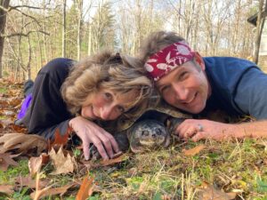 A photograph of Sy Montgomery and Matt Patterson, laying on their stomachs in grass and fallen leaves. Nestled between their faces is Fire Chief, a snapping turtle saved by Turtle Rescue League and rehabilitated by Sy and Matt.