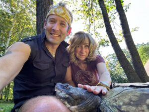 A selfie photograph taken by Matt Patterson. He and Sy Montgomery sit on the ground beneath trees, and Fire Chief, a common snapping turtle saved by Turtle Rescue League and rehabilitated by Matt and Sy, sits with them, with his chin resting on Matt's leg. Sy pats the top of his head.